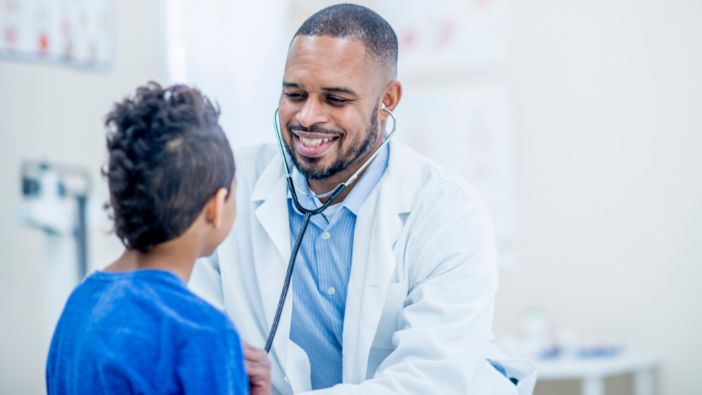 The Waiting Room Blog 1 Smiling male doctor using a stethoscope to examine a young boy in a medical office.