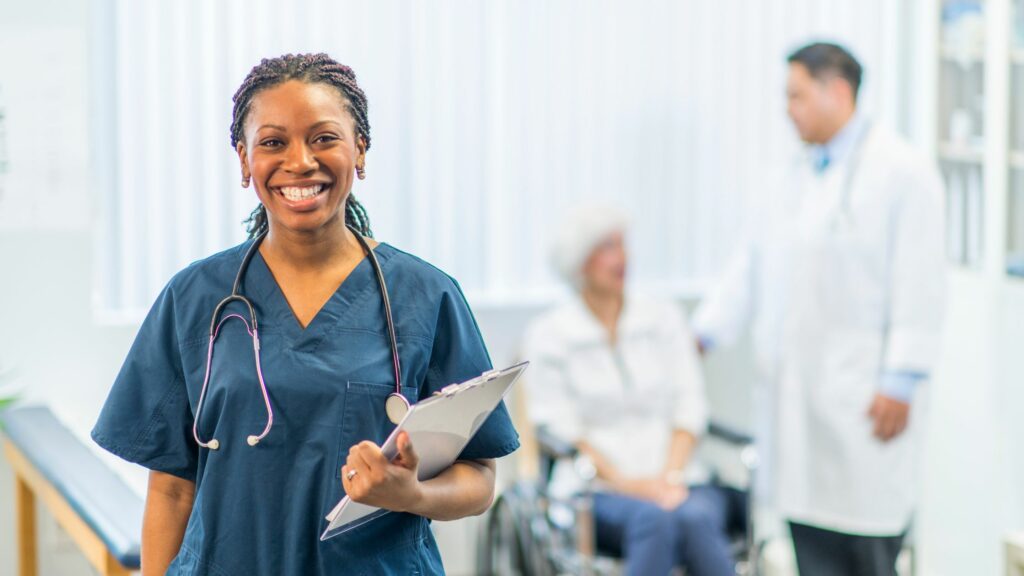 Nurse standing with stethoscope around neck holding a clipboard. Blurred in the background is a doctor and elderly patient in a wheelchair.