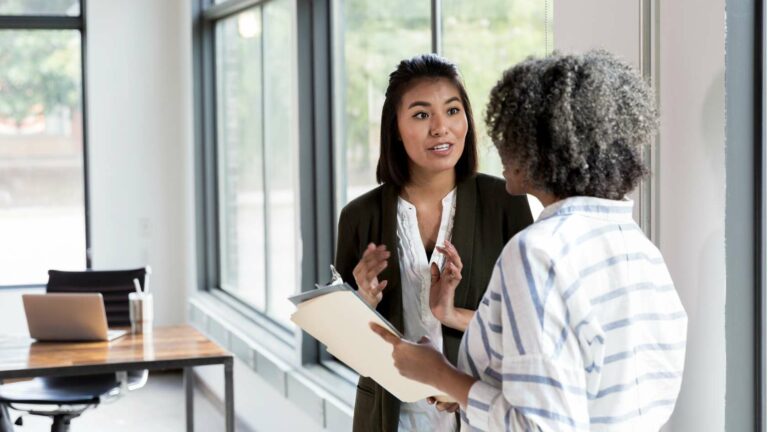 Two women in an office setting, speaking to each other, one with a clipboard in hand.