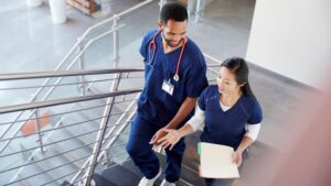 Two nurses in scrumbs walking up a staircase. One holding a clipboard with paperwork.