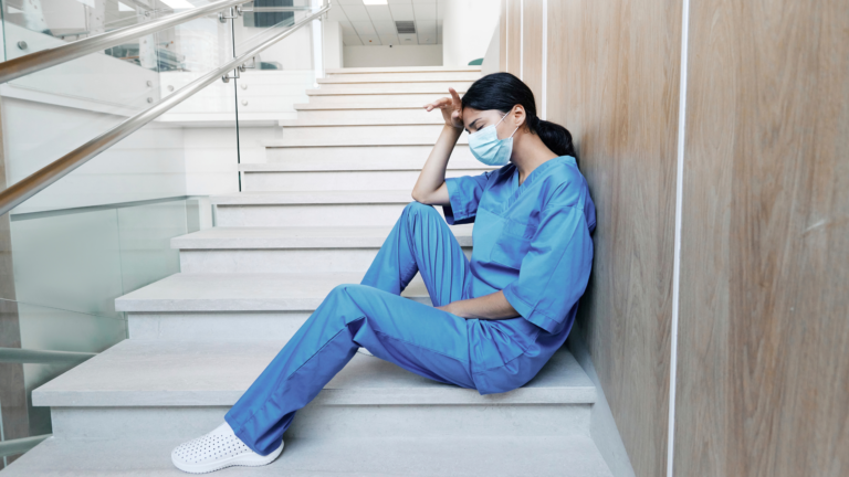 Tired nurse in scrubs sitting on steps with hand on forehead.