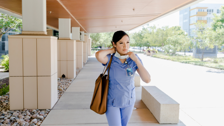 Female nurse walking outside a hospital, with side bag, adjusting face mask.